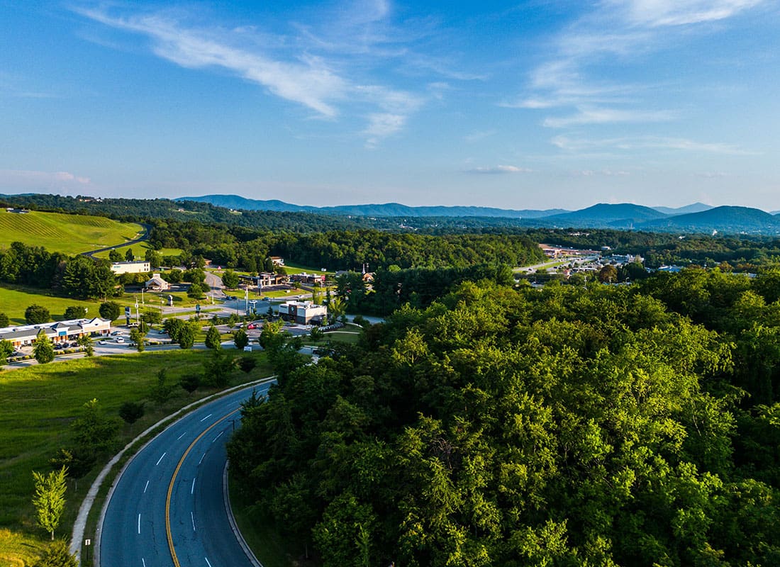 Insurance Solutions - Aerial View of an Empty Road Going Through a Small Town Surrounded by Green Trees with Mountains in the Distance Against a Blue Sky
